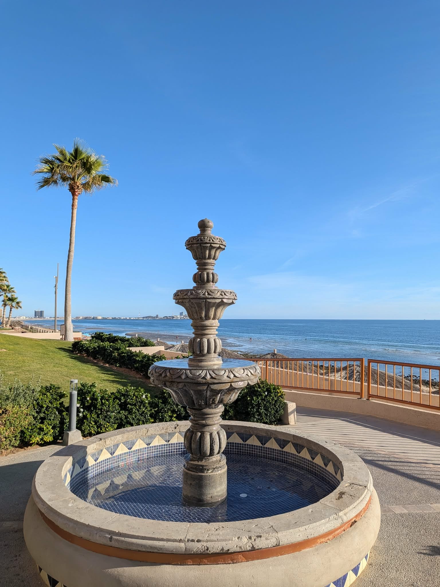 Tiered stone fountain on the resort terrace with a palm tree and the sea beyond
